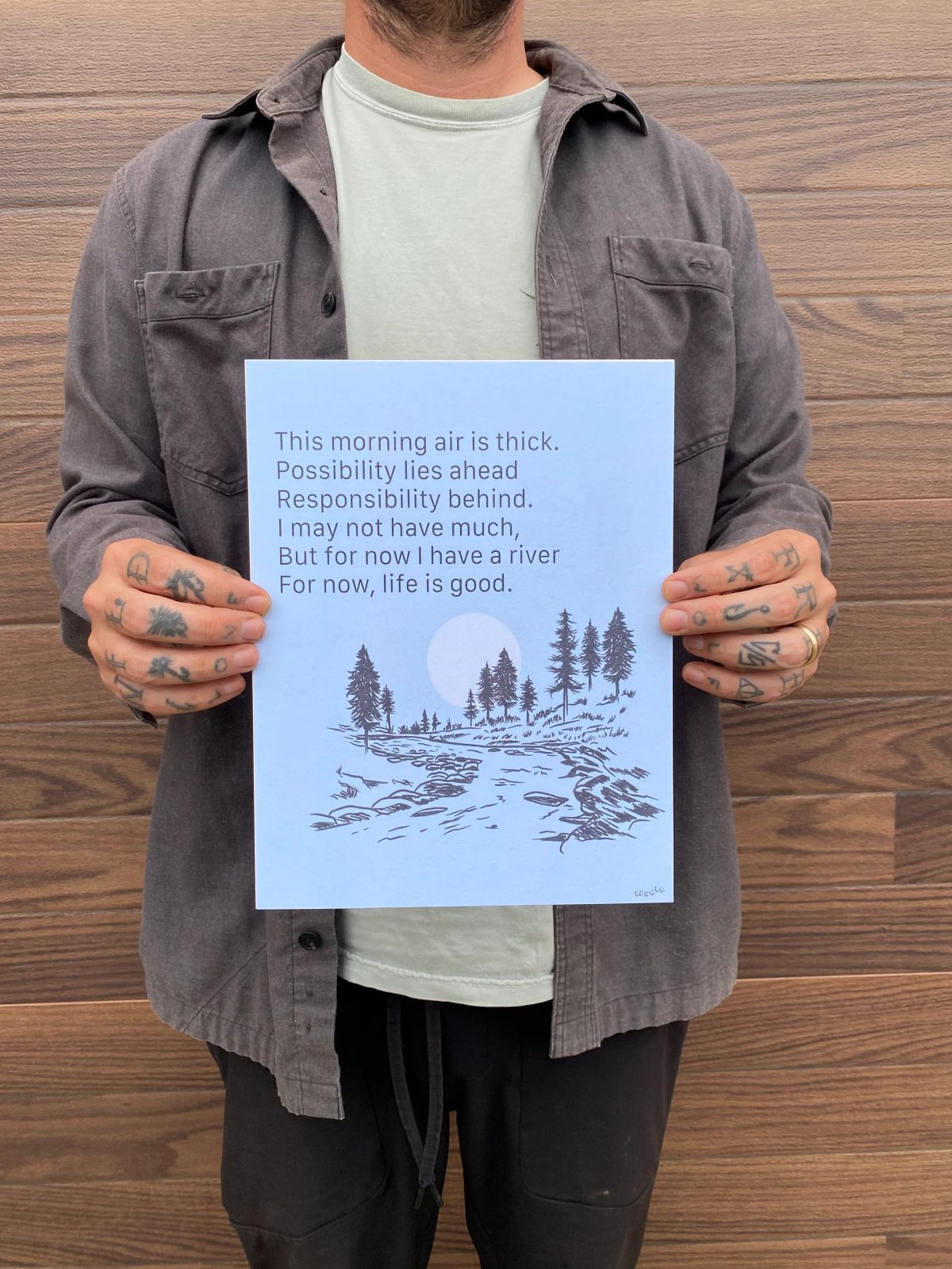 the artist holding a print with text and a river running by trees, the text reads: 